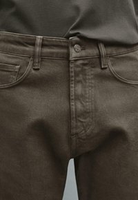 Close-up of dark brown denim pants waist with button fastened and part of a tucked-in dark shirt visible.