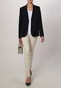 Blazer marine à deux boutons, T-shirt blanc, pantalon beige clair ajusté et escarpins noirs. Tient un petit sac à main noir avec des accents dorés.