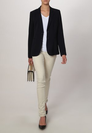 Blazer blu navy con due bottoni, t-shirt bianca, pantaloni fitted beige chiaro e tacchi alti neri. Tiene una piccola borsa nera con dettagli dorati.