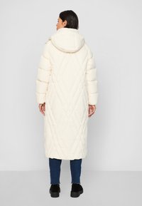 Long, quilted coat in cream with a hood, featuring a diamond pattern and side zippers. Paired with dark jeans and black shoes.