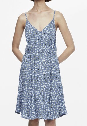 Vrouw die een blauwe mouwloze jurk met witte bloemenprint en dunne schouderbandjes draagt, staand met haar handen achter haar rug.