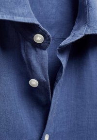 Chemise en lin bleu avec un col classique, des boutons blancs, et un tissu texturé. Vue en gros plan mettant en valeur les détails des boutons et du col.