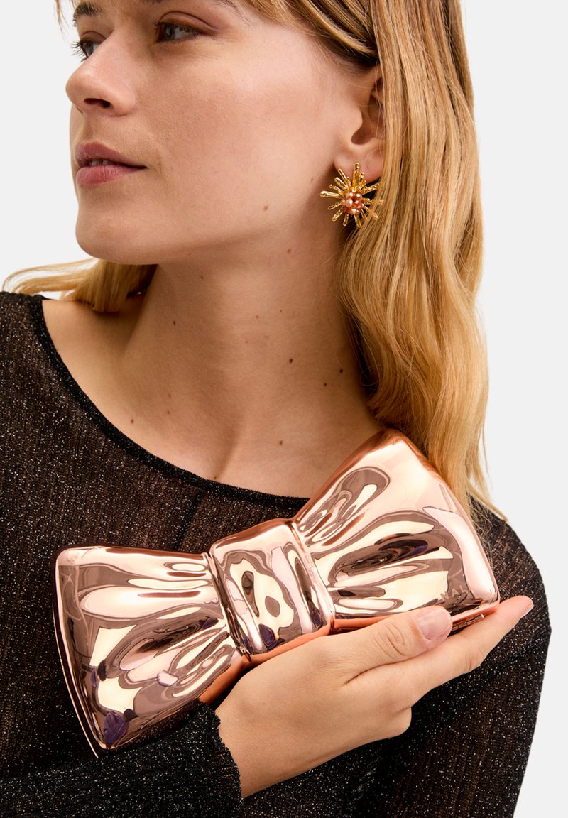 Metallic roséguldfärgad clutch med båtformat som hålls mot en svart glittrig topp, med en polerad yta och en upphöjd central knutdetalj.