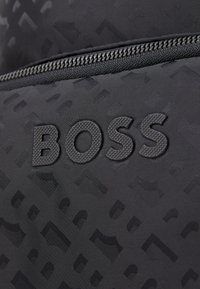 Fekete táska, amely texturált anyagból készült, diszkrét mintával. A "BOSS" logó matt fekete színben van gravírozva az elején. Zippzár részletek láthatók.