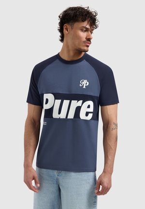 Mann mit lockigem Haar trägt ein blaues und marineblaues T-Shirt mit dem Schriftzug „Pure“ in großen weißen Buchstaben auf der Brust und hellblaue Jeans.