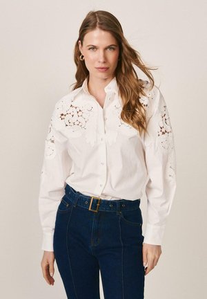 Mujer de cabello largo y castaño que lleva una blusa blanca abotonada con incrustaciones de encaje y jeans de talle alto azul oscuro con cinturón, sobre un fondo neutro.