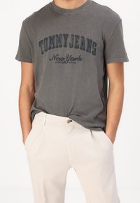 Šedé bavlněné tričko s logem "TOMMY JEANS New York" v tmavě modrém potisku, doplněné světle béžovými kalhotami. Klasický design s kulatým výstřihem.