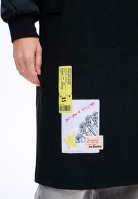 Ruka spočívá vedle černého kabátu s žlutou nášivkou ve formě lístku a bílou nášivkou zobrazující abstraktní tváře a nápis: "NEVYRŮSTĚJ. JE TO JÍZDA."