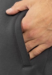 Šedá látková kapsa s viditelnou rukou, která se chytá za okraj, vystavující hladkou strukturu a detail šití nad černým akcentem.