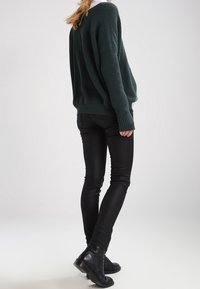 Suéter de punto de color verde oscuro y oversize combinado con jeans negros ajustados y botas de tobillo; textura de canalé visible y ajuste relajado.