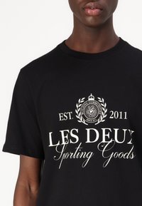 Juodas medvilninis marškinėlis su balta tekstu ir logotipu, kuriame parašyta "LES DEUX Sporting Goods" ir "EST. 2011," su klasikiniu apvaliu iškirpimu.