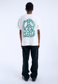 Bílý bavlněný tričko s grafikou zeleného květinového symbolu míru na zadní straně, v kombinaci s černými kalhotami a tmavými teniskami.