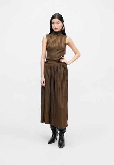 Χάλκινο μπλουζάκι χωρίς μανίκια με ρίγες, συνδυασμένο με μια αντίστοιχη πλισέ φούστα, που έχει απαλή υφή και μαύρες εφαρμοστές μπότες μέχρι το γόνατο.