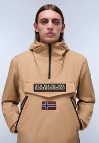 Beige hættetrøje med frontlynlås, med et stort logo-patch og et Norge-flag badge. Lavet af et tekstureret, vandafvisende materiale.