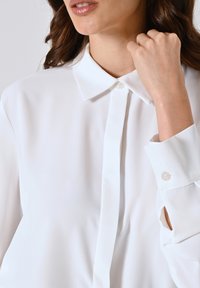 Blusa bianca a maniche lunghe con colletto a bottoni, tessuto morbido e fessure nei polsini, caratterizzata da chiusura anteriore con bottoni e linee pulite.