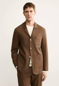 Blazer marrone con colletto a rever, due tasche a toppa e chiusura con bottoni. Indossato sopra una maglietta bianca a collo rotondo. Texture liscia, vestibilità su misura.