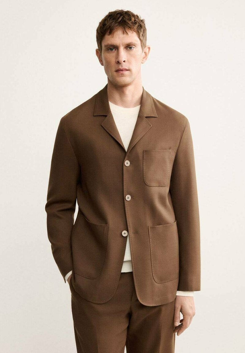 Blazer marrone con colletto a rever, due tasche a toppa e chiusura con bottoni. Indossato sopra una maglietta bianca a collo rotondo. Texture liscia, vestibilità su misura.