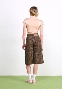 Person standing on green floor wearing beige cropped top, leopard print knee-length shorts, white socks, and beige sneakers, viewed from behind.
