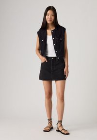 Νεαρή γυναίκα με μακριά σκούρα μαλλιά, ντυμένη με αμάνικο μαύρο τζιν γιλέκο, λευκό τοπ, μαύρη φούστα από τζιν και μαύρα σανδάλια με δέσιμο.