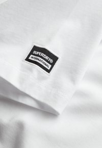 Hvid bomulds T-shirt med et sort rektangulært mærke med "SUPERDRY" og "INTERNATIONAL" i hvid tekst. Glat stof og enkelt design.