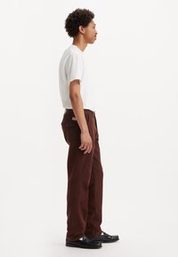 Pantaloni maro evazați cu buzunare în față și în spate, asortați cu un tricou alb simplu și mocasini negri, văzuți din lateral.