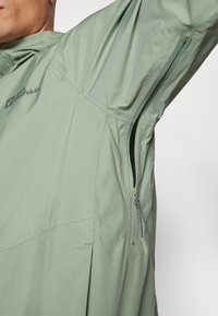 Chaqueta ligera de color verde claro que presenta un bolsillo lateral con cremallera, tejido texturizado y un logo en el pecho para un uso práctico al aire libre.