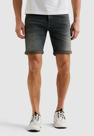 Muž v tmavých džínových kraťasoch s vyhrnutými nohavicami, bielych teniskách a bielom tričku stojaci pred jednoduchým svetlým pozadím.