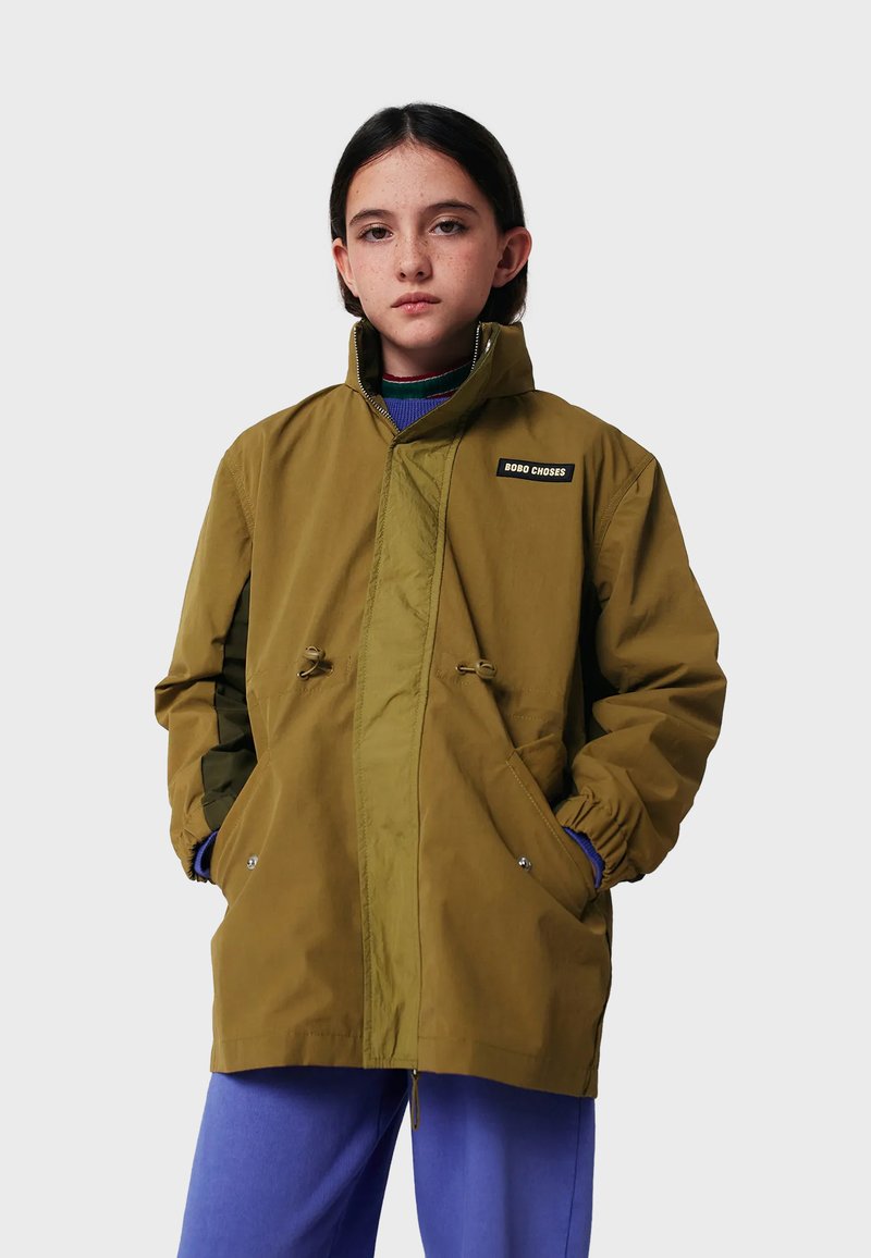 Junge Person mit dunklem Haar trägt eine olivegrüne Jacke mit Reißverschluss, hat die Hände in den Taschen und trägt blaue Hosen vor einem schlichten grauen Hintergrund.
