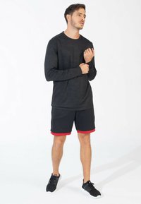 Sort langærmet træningsshirt med struktureret mønster, parret med sorte shorts med et rødt strib. Modellen bærer sorte træningssko.