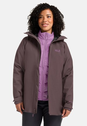 Donna con capelli ricci che indossa una giacca esterna marrone sopra una giacca viola con zip, in piedi davanti a uno sfondo chiaro uniforme.