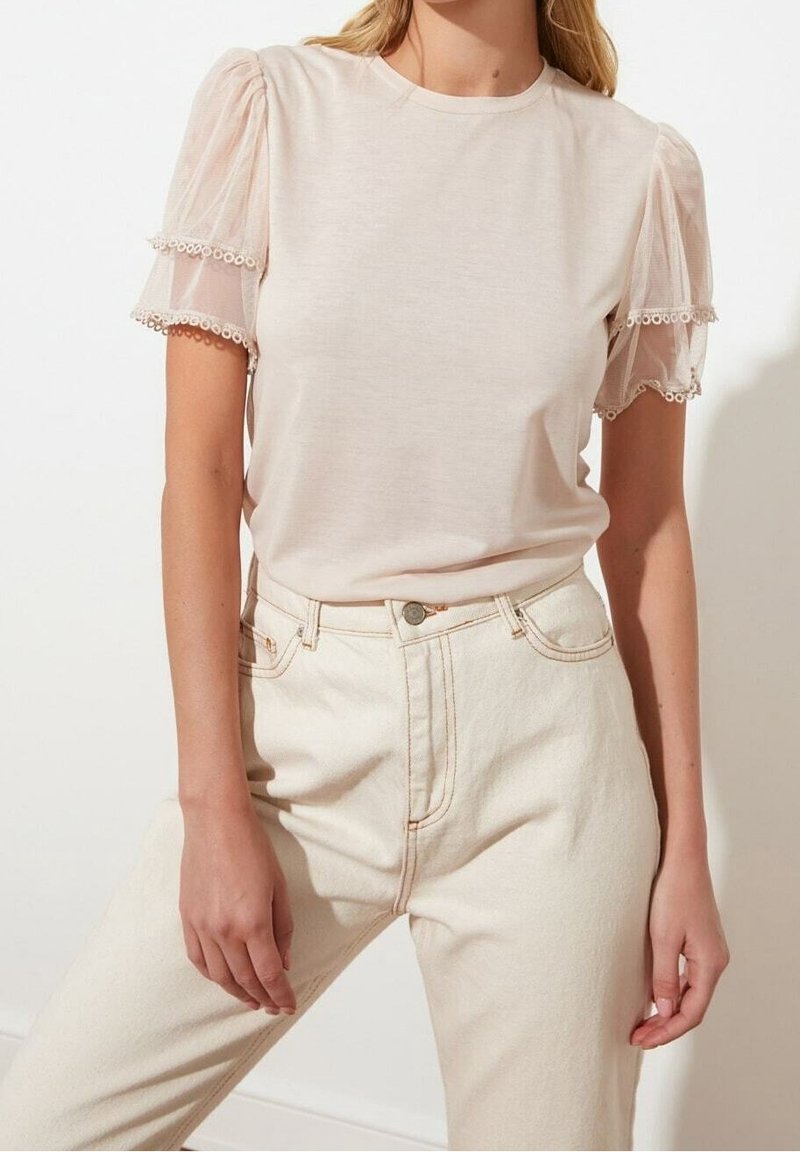 Maglietta rosa chiaro con maniche a sbuffo in tessuto trasparente e rifiniture in pizzo, abbinata a jeans beige a vita alta, che mettono in risalto uno stile pulito e minimalista.