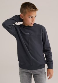 Mladý chlapec s krátkymi blond vlasmi, oblečený v tmavozelenej mikine s nápisom "Blue Ridge Denim" a svetlo šedých džínoch, pózujúci s rukou za hlavou.