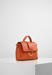 Sac à main en cuir orange à la forme structurée, avec une fermeture à rabat, un accent de quincaillerie ronde et des clous dorés le long des côtés.