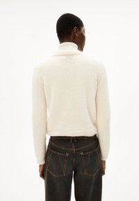 Maglione di lana color crema con colletto alto, lavorazione a maglia testurizzata e orlo a coste, abbinato a jeans di denim scuro con doppie tasche.