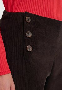 Pantalon en velours côtelé marron avec une taille ajustée et trois boutons décoratifs sur le côté. Le tissu a une apparence texturée et côtelée.