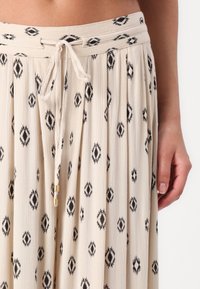 Jupe plissée en beige clair avec des motifs géométriques noirs. Dotée d'une taille à cordon de serrage avec un accent doré et d'un tissu doux et texturé.