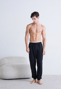 Schwarze Jogginghosen mit elastischem Bund und Kordelzug. Weiches, leichtes Material mit lockerer Passform. Barfuß männliches Modell steht.