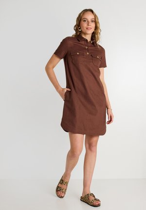 Vestido corto de lino marrón con cuello, dos bolsillos frontales, botones dorados y un corte relajado. Combinado con sandalias de estampado de leopardo.