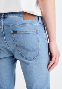 Jeans de mezclilla de color azul claro con un diseño clásico de bolsillo trasero, con costuras en contraste y una etiqueta de cuero marrón en la cinturilla.