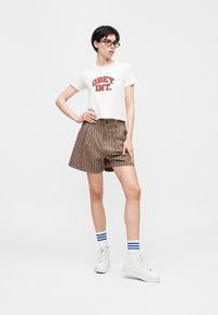 Γυναίκα με γυαλιά, λευκό κοντό t-shirt με το λογότυπο "OBEY INT.", καφέ καρό σορτς, λευκές κάλτσες με μπλε ρίγες και λευκά platform sneakers, στέκεται με το χέρι στη μέση.