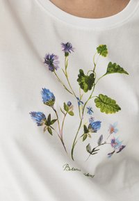 Bílá bavlněná tričko s botanickým potiskem modrých a fialových květin a zelených listů. Text "Botanické zahrady" v zelené barvě.