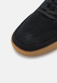 Sapatilha de tecido preta com um upper texturizado, apresentando uma biqueira arredondada e uma sola em borracha natural. Costura reforçada ao longo da borda.