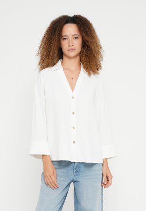 Λευκό πουκάμισο με κουμπιά, V-λαιμόκοψη, φαρδιά μανίκια και χρυσά κουμπιά. Το ύφασμα έχει υφή, σε συνδυασμό με μπλε τζιν παντελόνι.