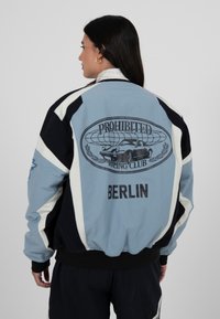 Femme aux longs cheveux noirs portant une veste bleue et noire avec les inscriptions « Prohibited Racing Club » et « Berlin » ainsi qu'un graphisme de voiture dans le dos.