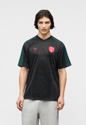 Nuori mies, jolla on hartioille ulottuvat tummat hiukset, pukeutuneena mustaan Adidas-paitaan, jossa on vihreitä raitoja ja punainen All-Stars 1894 -logo, yhdistettynä vaaleanharmaisiin Adidas-housuihin.