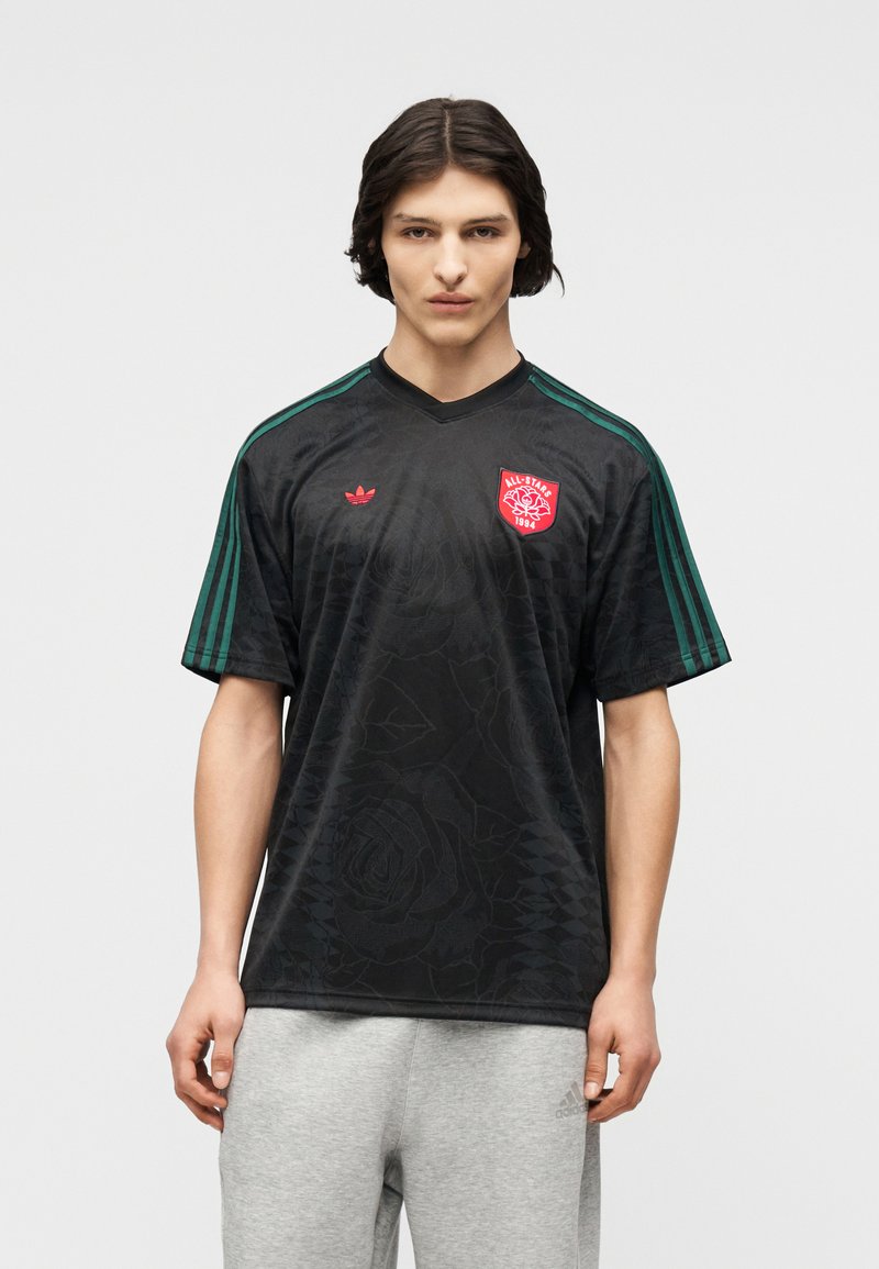 Νέος άνδρας με σκούρα μαλλιά μέχρι τους ώμους που φοράει μια μαύρη φανέλα Adidas με πράσινες ρίγες και κόκκινο λογότυπο All-Stars 1894, συνδυασμένη με ανοιχτό γκρι φόρμα Adidas.