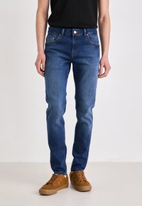 Jeans de mezclilla azul con un corte ajustado, que cuentan con cinco bolsillos, una cinturilla con trabillas para el cinturón y costuras a contraste. Combinados con zapatillas marrones.