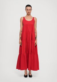 Abito lungo rosso senza maniche con volant a strati. Realizzato in tessuto leggero, presenta un colletto rotondo e una silhouette fluida. Scarpe nere indossate.