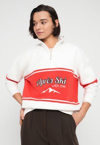 Λευκό και κόκκινο φούτερ με μισό φερμουάρ, με κείμενο "Après Ski with me" και γραφικό βουνού. Μοντέρνα μεγάλη εφαρμογή και ριμπ μανσέτες.