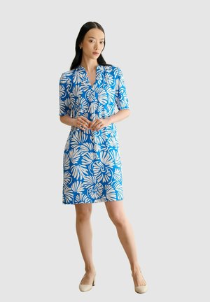 Mujer con vestido azul con estampado floral blanco, mangas a medio brazo, de pie con las manos entrelazadas, calzando tacones beige sobre fondo liso.
